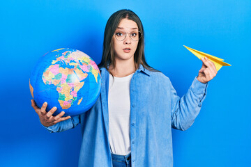 Fototapeta premium Young hispanic girl holding paper plane and world ball in shock face, looking skeptical and sarcastic, surprised with open mouth