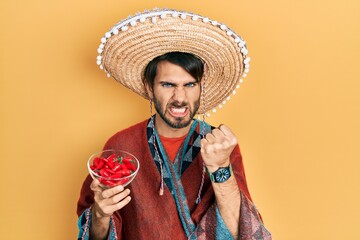 Young hispanic man wearing mexican hat holding chili annoyed and frustrated shouting with anger,...