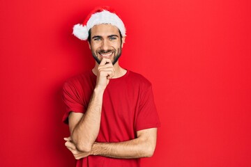 Young hispanic man wearing christmas hat looking confident at the camera with smile with crossed arms and hand raised on chin. thinking positive.