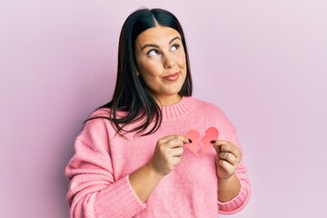 Beautiful brunette woman holding broken heart paper shape smiling looking to the side and staring away thinking.