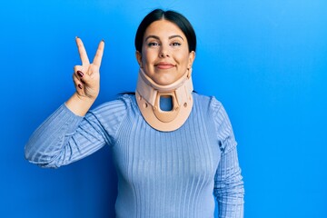 Beautiful brunette woman wearing cervical collar smiling with happy face winking at the camera doing victory sign. number two.