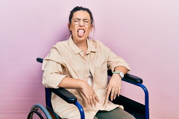 Young hispanic woman sitting on wheelchair sticking tongue out happy with funny expression. emotion...