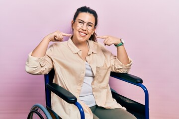Young hispanic woman sitting on wheelchair smiling cheerful showing and pointing with fingers teeth...