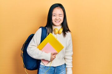 Young chinese girl holding student backpack and books winking looking at the camera with sexy...