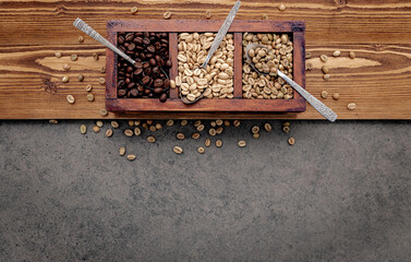 Green and brown unroasted and dark roasted coffee beans in wooden box with spoons setup on dark...