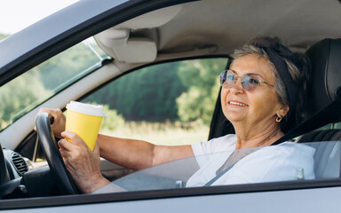 Portrait of a happy mature woman with glasses holding a disposable cup of coffee while driving a car and looking out the open window. Active smiling pensioner driver sitting at the wheel, outdoors.