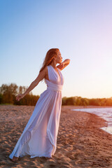Fototapeta na wymiar A young woman of European appearance in the summer in a white dress on the seashore stands at sunset. Enjoys loneliness and the sea breeze