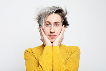 Portrait of funny shocked young man in yellow sweater holding cheeks standing on light grey background