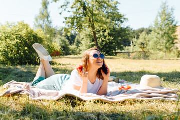 Happy young woman lying on a blanket and talking on a cell phone during a summer outdoor picnic....