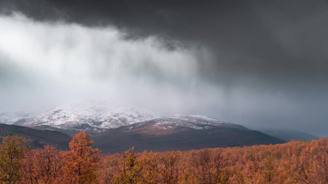 Snow and rain are falling above the autumn forest. Dark clouds moving swiftly over the snow-covered mountains. The storm is coming. Timelapse video.