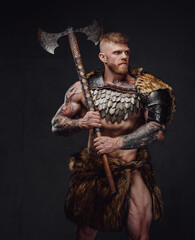 Brutal tattooed warrior wearing light armour and fur holding two-handed axe in dark studio