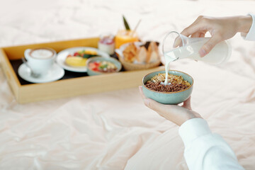 Hands of young woman adding milk in bowl of granola when sitting on bed with breakfast tray