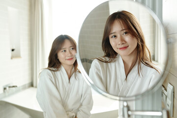Fototapeta premium Smiling attractive young woman in bathrobe looking at magnifying mirror after taking shower