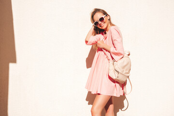 Young beautiful smiling hipster woman in trendy summer pink dress. Sexy carefree woman posing near white wall in the street at sunset. Positive model outdoors.Happy and cheerful