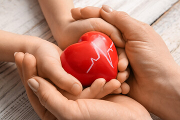 Hands of woman and child with red heart on light wooden background, closeup