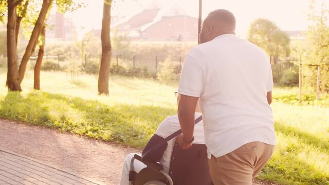 African-American caregiver and old disabled man in a wheelchair. Professional nurse and handicapped patient in the park. Assistance, rehabilitation and health care. Golden hour sunset.