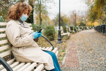 Fototapeta na wymiar Corona communication. Covid leisure. Quarantine lifestyle. Curly hair overweight woman in medical face mask outside phone chatting in autumn park landscape copy space.