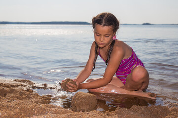 Fototapeta na wymiar A tanned girl is building a house of sand on the shore of the lake.