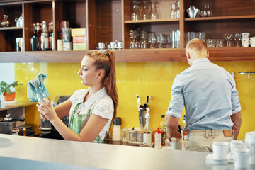 Serious young barista wiping glass jars with soft cloth when manager preparing drinks ingredients in background