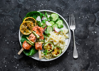 Salmon skewers with zucchini, pasta and vegetables salad on a dark background, top view. Delicious healthy balanced lunch
