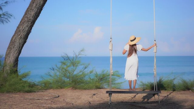 With her back to the camera, a young woman in a white sundress and straw sun hat stands on a large rope swing to get a better view of the ocean in the distance. Title space