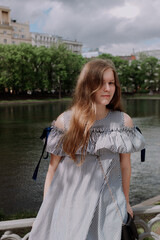 Smiling young woman in dress standing alone among street, leaning on fence in front of pond. looking at camera. Having fun outside. Urban style and street fashion. Sumeer casual outfit