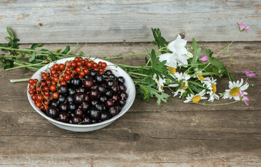 A plate with black and red currants next to a delicate bouquet of meadow flowers: chamomiles and bells on the wooden steps of an old house. Summer holidays concept. Still life. Summer time