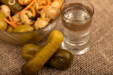 A glass of vodka, pickled cucumbers and salted mushrooms in a glass dish on a background of homespun fabric with a rough texture. Close-up Selective focus.