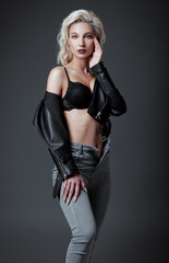 Studio fashion: beautiful young woman in black leather jacket, bra and jeans. Seductive informal (rock) girl against gray background