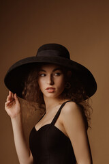 Fototapeta na wymiar Fashion portrait of elegant curly brunette woman wearing luxury wide brim hat, black dress. Model looking aside
