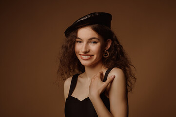 Beautiful happy smiling fashionable curly brunette woman wearing trendy leather beret, golden earrings, stylish black dress, posing in studio on brown background. Copy, empty space for text
