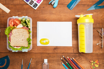 A lunch box with a sandwich, vegetables, water, fruit and a notebook on a wooden background. School...