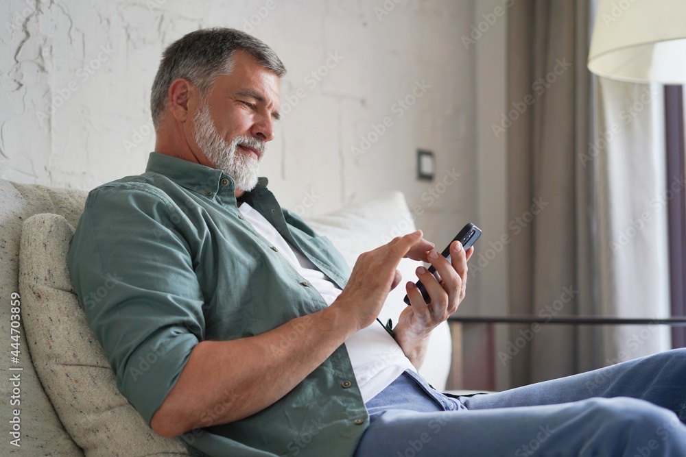 Wall mural smiling senior man using cellphone browsing internet sitting on sofa indoor.