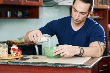 Fit muscular man adding fresh milk in bowl of granola when eating healthy breakfast in kitchen