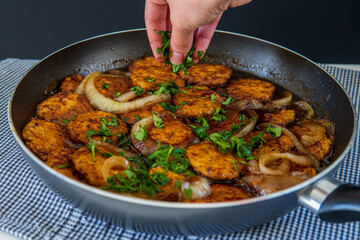 Woman's hand sprinkles parsley over sweet and sour chicken patties in a pan, on a plaid towel. Chicken cutlets cooked with Asian flavored sauce and caramelized onions. Chinese style dish, side view.