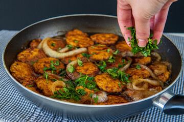 Woman's hand sprinkles parsley over sweet and sour chicken patties in a pan, on a plaid towel. Chicken cutlets cooked with Asian flavored sauce and caramelized onions. Chinese style dish, side view.