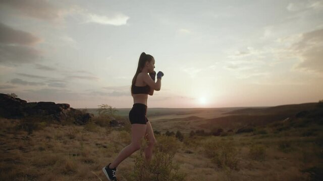 Sport Woman with bandages on her hands trains boxing punches with moving forward in nature against background of sunset. Girl motivation trains to box in outdoor