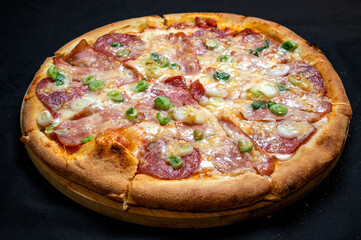 Pizza with green onions, salami, ham, mozzarella and tomato sauce on a black background.