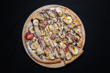 Pizza with chocolate sauce, caramel sauce, strawberries, bananas, pineapple and powdered sugar on a black background.
