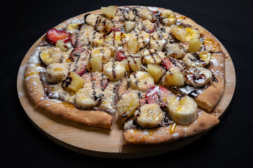 Pizza with chocolate sauce, caramel sauce, strawberries, bananas, pineapple and powdered sugar on a black background.