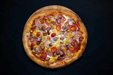 Pizza with tomato sauce, mozzarella, sour cream, salami, pork ham, sausages, corn and cherry tomatoes on a black background
