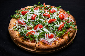 Pizza with tomato sauce, smoked pork muscles, mozzarella, arugula salad and cherry tomatoes on a black background.