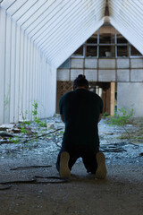 Man in dark clothes kneeling in despair in destroyed church building, praying, back view