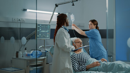 Nurse and african american doctor treat sick patient in hospital ward with modern medical equipment. Old man with nasal oxygen tube and oximeter waiting for treatment against disease