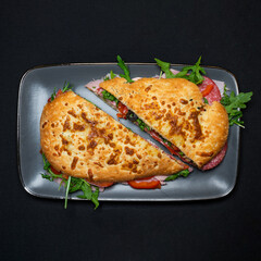 sandwich with salami, pork ham, peppers, olives and arugula salad on a black background.