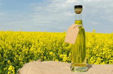 Glass bottle full of canola oil and paper tag for text or price against canola field and blue sky