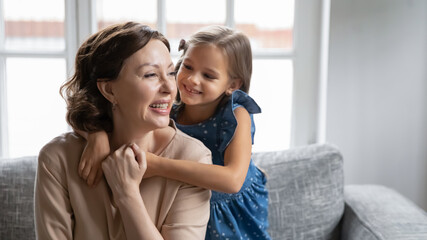 Fototapeta premium Cute little Caucasian teen girl child hug cuddle smiling old grandmother show love and care to grandparent. Happy small granddaughter embrace middle-aged granny relax rest together on sofa at home.
