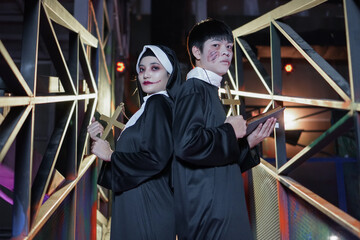Asian young woman and man dressed as nun and priest holding the cross and holy book in hands and make up face for blood and wounds at Halloween party. Dark background. Silhouette