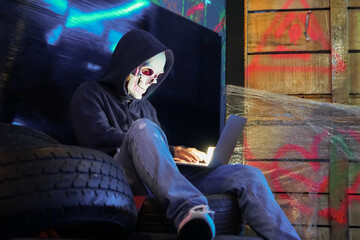 Anonymous man in hoodie and pink neon skull mask hacking into laptop computer and stealing personal...