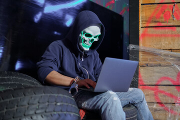 Anonymous man in hoodie, shackle and green neon skull mask hacking into laptop computer and...